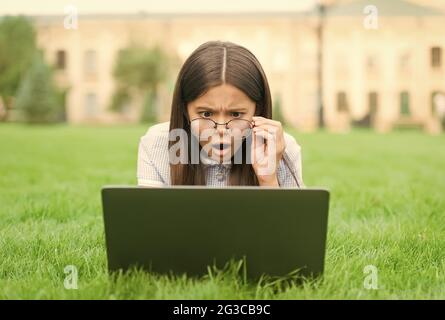 Oh non. Retour à l'école. Jeune fille utiliser l'ordinateur sur l'herbe verte dans le parc. Enfant dans les lunettes avec le carnet. Nouvelle technologie dans la vie moderne. Étude. Surpris Banque D'Images