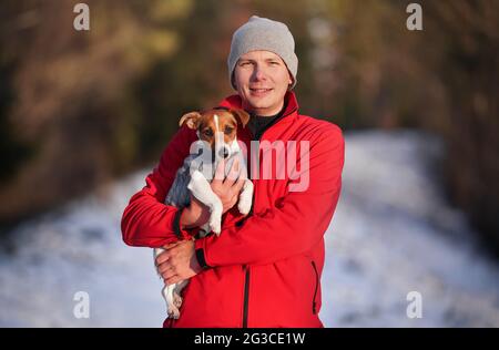 Jeune homme en blouson rouge portant son chien terrier Jack Russell à la main l'après-midi d'hiver, neige floue sur fond de sentier couvert Banque D'Images