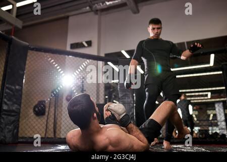 Un homme d'artiste martial mixte en noir gagne, se tenir au-dessus du combattant perdant après des combats durs dans la bague à la salle de gym. Boxe, MMA, sport concept. Concentrez-vous sur le shi Banque D'Images