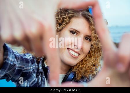 Gros plan portrait d'une jeune heureuse cheveux bouclés femme caucasienne regardant à travers un cadre fait de ses doigts. Capturez le concept du moment. Banque D'Images