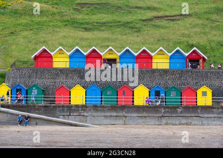 Les gens apprécient les plaisirs de la mer britannique en face d'une rangée de cabanes de plage colorées sur la plage de Whitby dans le North Yorkshire, comme plus de gens décident Banque D'Images