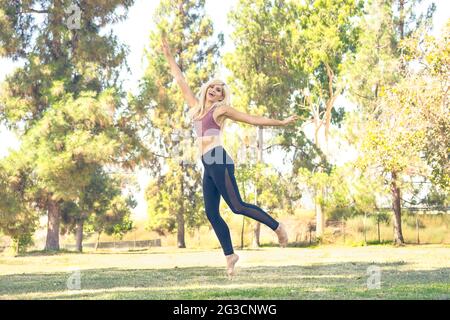 Une femme de race blanche qui saute, portant une tenue de yoga se posant à l'extérieur dans un parc entouré d'arbres. Banque D'Images