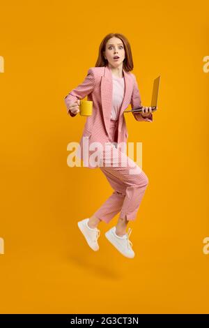 REDHEAD femme saut en utilisant ordinateur portable web projet isolé sur fond jaune en studio, tenant une tasse de café en mains, manquer la date limite, hur Banque D'Images