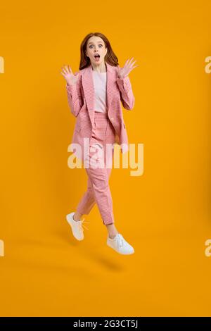 Photo pleine grandeur de jeune heureux surpris surpris surpris la femme sautant dans l'excitation isolée sur fond jaune, portant le costume rose, élevant ha Banque D'Images