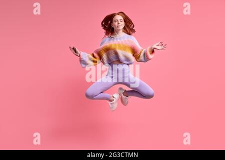 belle dame à tête rouge sautant de l'air assis haut dans lotus pose pratiquant le yoga du matin portant une tenue décontractée isolé pastel couleur rose fond, por Banque D'Images