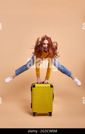 Voyageur excité touriste redhead femme avec valise, sautant dans une tenue décontractée isolée en studio. Passager caucasien voyageant à l'étranger la semaine Banque D'Images