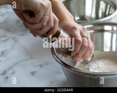Gros plan sur les mains d'une femme tenant un moulin à sel sur un bol de mélange en métal sur une surface en marbre avec lumière naturelle Banque D'Images
