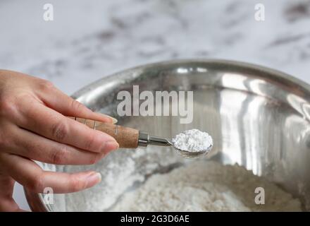 tenir à la main une cuillère de mesure avec de la poudre blanche dans un saladier rond en métal rempli d'autres ingrédients Banque D'Images