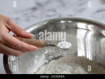 main de femme tenant une cuillère de mesure avec de la poudre blanche dans un bol de mélange rond en métal rempli d'autres ingrédients Banque D'Images