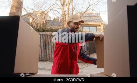 Le liveur porte un capuchon et des boîtes en carton de levage uniformes rouges. Pile de boîtes à colis. Concept de livraison de marchandises d'expédition de service. Employé du courrier. Photo de haute qualité. Banque D'Images