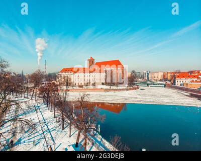 Wroclaw, Pologne 02.15.2021 - Paysage couvert de neige avec des arbres toujours verts sur le côté d'une rivière. Zone industrielle avec cheminées de centrales thermiques. De la fumée s'échappe et pollue le ciel bleu clair. Banque D'Images