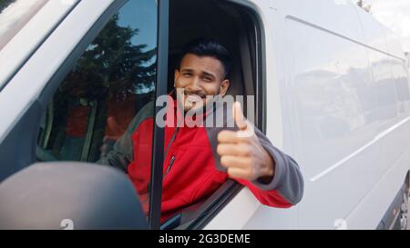 Un préposé à la livraison portant un uniforme rouge assis dans la camionnette blanche faisant un signe de pouce vers le haut. Joyeux Indien assis sur le siège avant et souriant à la caméra. Concept de messagerie. Banque D'Images
