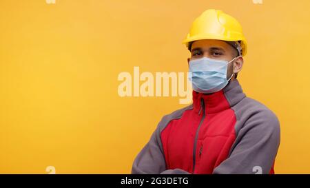 Portrait d'un jeune ingénieur indien portant un casque jaune de sécurité et un masque debout avec les bras croisés dans le studio avec fond jaune. Homme isolé. Gros plan. Banque D'Images