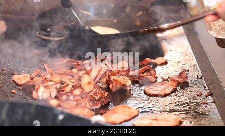 Gros plan de beaucoup de bacon croustillant cuit sur une plaque de barbecue chaude avec une cuisson à la crêpe dans une casserole en arrière-plan. Banque D'Images