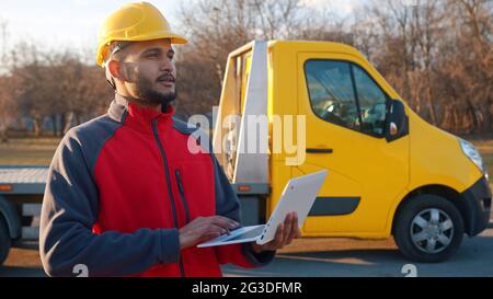 Un jeune ingénieur portant un uniforme et un casque de sécurité debout avec un camion jaune derrière lui. Tenir un ordinateur portable dans la main. Concept d'ingénierie et de construction. Banque D'Images