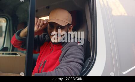 Un livreur portant un uniforme rouge assis dans la camionnette blanche et renverse sa casquette. Joyeux Indien assis sur le siège avant et souriant à la caméra. Concept de livraison de colis. Banque D'Images