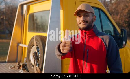 Un employé portant un chapeau et un uniforme avec un camion jaune derrière lui. Garçon souriant et faisant un signe de pouce vers le haut. Concept de mécanicien automobile. La roue de secours est visible dans le chariot derrière. Banque D'Images