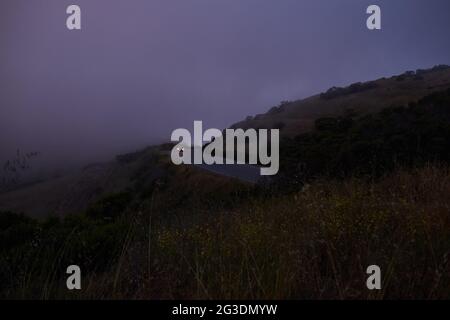 Vue de la route venteuse à travers les collines avec du brouillard et une voiture avec des phares conduisant dans la distance Banque D'Images