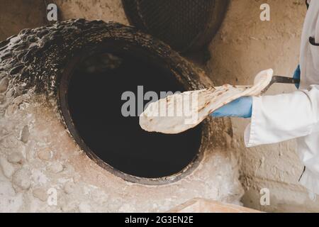 un homme âgé fait du pain traditionnel dans un vieux four rond en pierre dans un village rural. Cuisine d'Asie centrale. Pain chaud. Banque D'Images