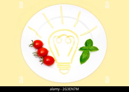 Composition créative des aliments : vue de dessus d'une assiette blanche avec un symbole d'ampoule lumineuse à base de pâtes cuites, de tomates cerises et de feuilles de basilic isolées Banque D'Images