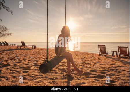 Jeune femme asiatique se détendant sur une balançoire en bois sur la plage au lever du soleil à la mer tropicale Banque D'Images