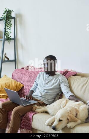 Homme africain en lunettes assis sur un canapé avec chien et travaillant en ligne à la maison Banque D'Images