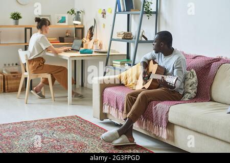 Homme africain jouant de la guitare sur un canapé avec une femme assise à la table et travaillant sur un ordinateur portable dans la chambre à la maison Banque D'Images