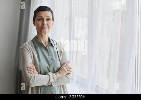 Portrait à la taille de la femme mûre regardant l'appareil photo tout en se tenant par la fenêtre avec les bras croisés, espace de copie Banque D'Images