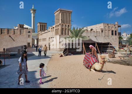 Une mère et une petite fille regardant un chameau et propriétaire de chameau dans le vieux quartier de Dubaï, les Émirats Arabes Unis., avec une maison avec une tour de vent de refroidissement juste au-delà Banque D'Images
