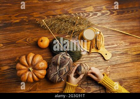 Mains de femmes avec une boule de fil, de citrouille, de café et de roseaux sur fond de bois. Mains dans un chandail orange. Artisanat, passe-temps, vue de dessus. Aiguilles de tricotage en bois en boule de fil. Bâtonnets de cacao et de cannelle. Banque D'Images