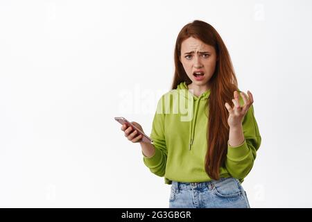 Frustré redhead fille se plaint à quelque chose sur le téléphone mobile, semble ennuyé et tenant smartphone, frowning déçu, fond blanc Banque D'Images