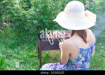 Une fille dans un chapeau de paille prépare de la viande fraîche aromatique sur le gril sur un fond de feuillage vert lors d'une journée d'été lumineuse. Gros plan Banque D'Images