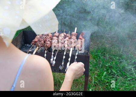 Une fille dans un chapeau de paille prépare de la viande fraîche aromatique sur le gril sur un fond de feuillage vert lors d'une journée d'été lumineuse. Gros plan Banque D'Images