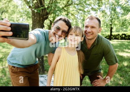 Couple gay faisant le portrait de selfie sur téléphone portable avec leur fille adoptée à l'extérieur Banque D'Images