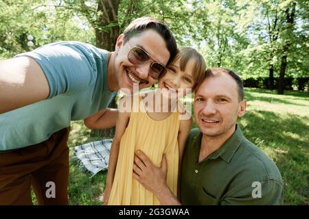 Portrait d'un couple de hommes gay heureux embrassant avec leur fille adoptée et souriant à l'extérieur de l'appareil photo Banque D'Images