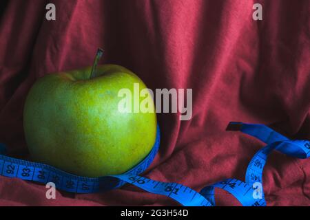 Une pomme verte sur fond de tissu rouge est enveloppée d'un ruban de mesure bleu. Le concept d'une nutrition adéquate et d'un mode de vie sain. Banque D'Images