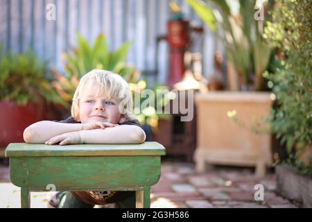 Joyeux adorable gamin australien blond assis et assis sur une petite table d'école dans la cour de la maison Banque D'Images