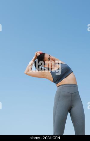 Photo vers le haut d'une jeune femme brunette dans les vêtements de sport gris faisant ses étirements. Un jour ensoleillé sous un ciel bleu clair. Banque D'Images