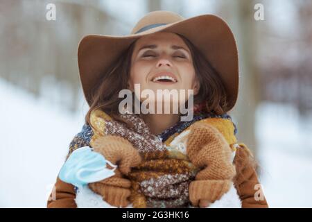 décontractée et élégante femme de 40 ans en chapeau marron et écharpe avec moufles et masque médical dehors dans le parc de la ville en hiver. Banque D'Images