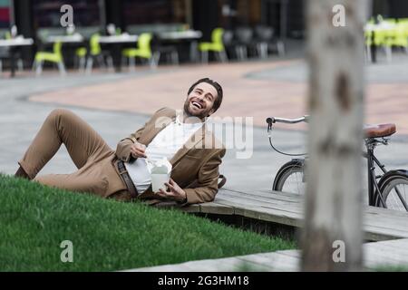 homme d'affaires heureux en costume tenant une boîte en carton avec des aliments asiatiques et des baguettes tout en étant couché près de l'herbe Banque D'Images