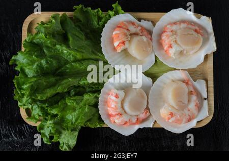 Un pétoncle brut naturel avec une queue de crevettes dans la coquille avec une feuille de laitue dans un bol en bois sur fond sombre. Mise au point sélective. Banque D'Images