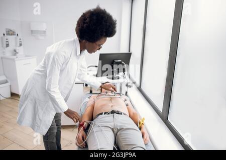 Test ECG du jeune homme à la clinique moderne. Jeune femme africaine médecin cardiologue mettant un équipement spécial, les électrodes de capteur de vide sur la poitrine du patient caucasien mâle avant de faire l'ecg Banque D'Images