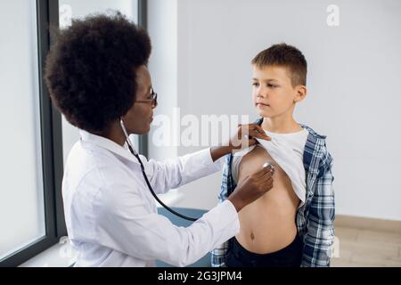 Pédiatre féminin africain avec stéthoscope ausculting mignon adolescent caucasien garçon patient. Médecin noir, vérifie le rythme cardiaque et les poumons de la respiration de l'enfant, faisant un contrôle avant la vaccination Banque D'Images