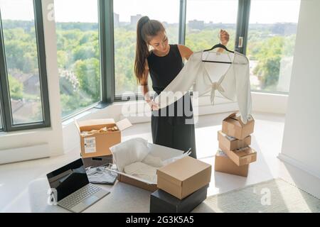 Vendre des vêtements en ligne en utilisant un ordinateur portable à la maison femme emballant de nouveaux vêtements dans des boîtes à expédier au client. Fille asiatique heureux de regarder le haut blanc à manches longues Banque D'Images