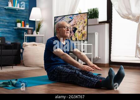 Pensioner focalisé portant des vêtements de sport assis sur le tapis de yoga étirement des jambes muscle pendant l'entraînement d'aérobic exercice musculaire résistance. Homme âgé faisant de l'exercice physique minceur poids Banque D'Images