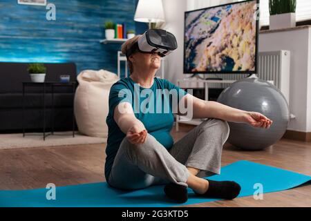 Femme sénior en bonne santé utilisant un casque de réalité virtuelle méditant tout en étant assise en position lotus sur le tapis de yoga dans le salon pendant l'entraînement de méditation. Pensioner entraînement corps muscle faire des sports exercice Banque D'Images