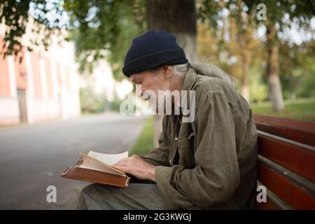 Side view of old homeless man with long grey hair reading a book. Stock Photo