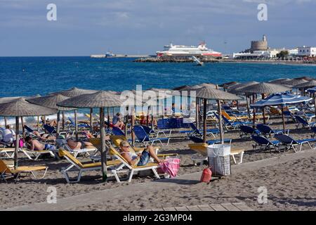 Les gens se reposant sur la plage, contre la baie et le port de la ville de Rhodes, l'île de Rhodes, Dodécanèse, Grèce Banque D'Images