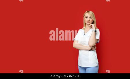 Médecin femme confiante debout sur fond rouge. Jeune femme stagiaire dans des vêtements médicaux posant. Banque D'Images