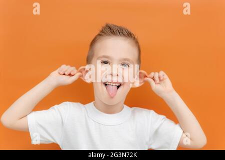 Un enfant fait un visage sur fond orange. Un garçon gai caucasien montre sa langue et tire ses oreilles. Un garçon dans un T-shirt blanc est crocheté Banque D'Images